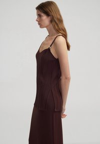 Abito a costine marrone con corpetto aderente, spalline sottili e un accento decorativo dorato sulla spalla. Il dettaglio con spacco laterale esalta la silhouette.