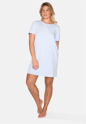 Jonge vrouw staat blootsvoets en draagt een lichtblauwe casual jurk met korte mouwen en een ronde hals op een effen achtergrond.