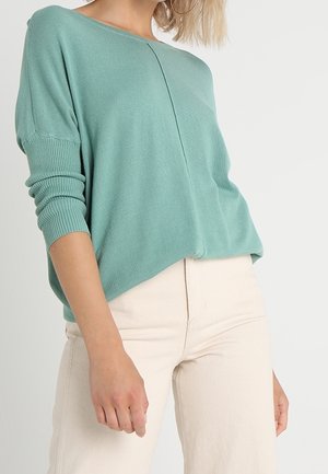 Maglione turchese a maglia con maniche lunghe e arrotolate, scollatura rotonda e vestibilità comoda, abbinato a pantaloni beige chiaro. Texture liscia.