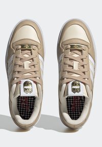 Pari bežových a bielych tenisiek Adidas so zlatými akcentmi, bielymi pruhmi po stranách a kockovanými vnútornými solami zobrazené zhora.