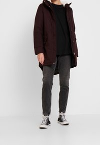 Giacca imbottita color bordeaux con cappuccio, foderata di morbido tessuto bianco, chiusura a zip e tasche laterali, abbinata a jeans scuri e sneaker nere.