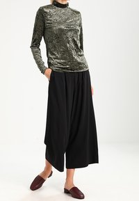 Olivgrünes, zerknittertes Langarmoberteil kombiniert mit schwarzen, weiten Hosen. Braune Slipper runden das Outfit ab und sorgen für einen lässigen Sitz.