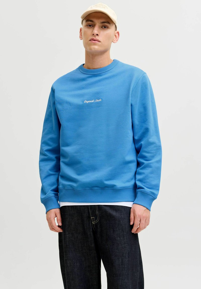 Mėlynas džemperis, pagamintas iš minkštos medžiagos, su apvaliu iškirpimu ir raštuotomis rankovėmis. Ant krūtinės yra balta siuvinėta užrašas.