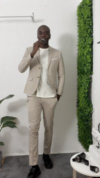 Bežový oblek s priliehavým strihom, biela košeľa pod ním a čierne topánky. Model drží malý predmet, v pozadí umelá zeleň.