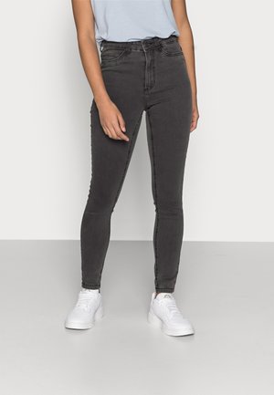 Personne portant un jean skinny taille haute gris foncé et des baskets blanches, debout devant un fond uni gris clair.