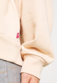 Felpa di colore crema con polsini a costina e un piccolo tag rosso di Levi's. La texture è morbida con una finitura liscia. Pantaloni grigi a quadri visibili.