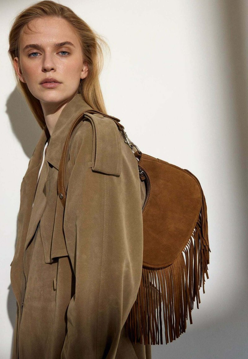 Tegen een tan kleurige suède trenchcoat met brede revers en epauletten, gecombineerd met een bruine suède schoudertas met franjes, die een ronde vorm heeft en uit waarschijnlijk zilveren hardware bestaat.