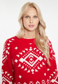 Pullover texturat roșu, cu un model geometric alb, având un guler rotund. Modelul are păr lung blond și poartă cercei colorati.