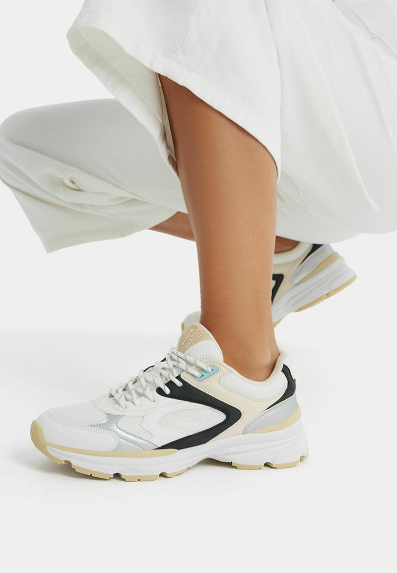 Weiße Mesh-Sportschuhe mit schwarzen und beigen Akzenten, strukturierten Paneelen, einem gepolsterten Kragen und einer dicken weißen Sohle mit einer hellbeigen Außensohle.