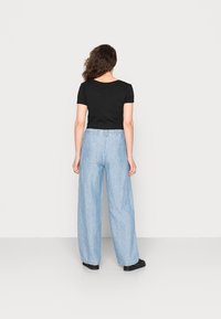 Vrouw met golvend bruin haar, gekleed in een zwarte korte mouwen shirt, lichtblauwe wijde broek en zwarte schoenen, staand tegen een witte achtergrond.