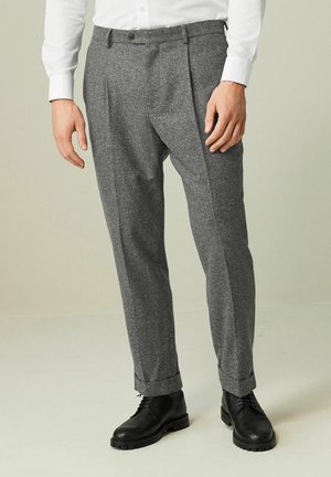 Homme portant un pantalon gris en chevrons avec revers, des bottes noires à lacets et une chemise blanche à manches longues boutonnée, debout devant un fond uni.