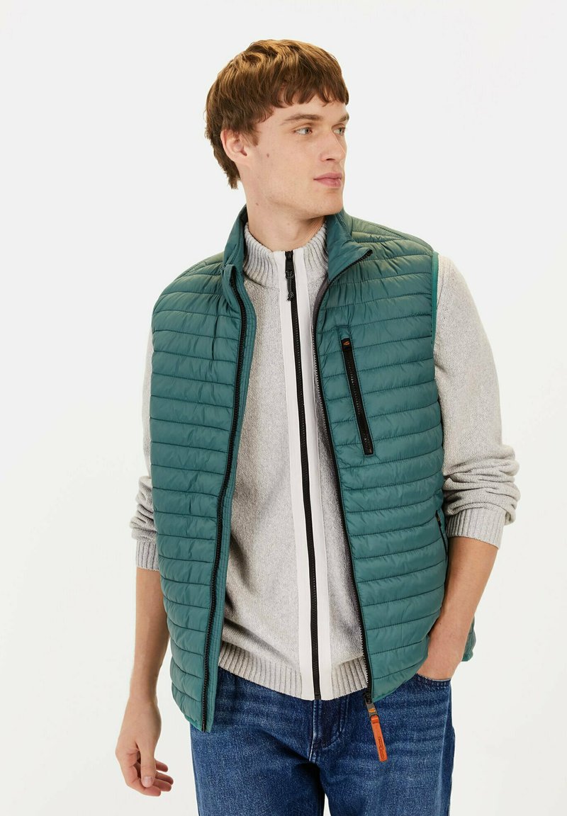 Gepolsterte türkisfarbene Weste mit Reißverschlussfront, ausgestattet mit einem Stehkragen und einer schwarzen Akzenttasche, über einem grauen Pullover und blauen Jeans getragen.