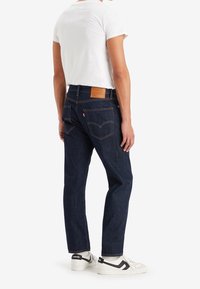 Jeans de mezclilla azul oscuro con corte recto, bolsillos traseros visibles y una etiqueta de cuero marrón en la cintura. Combinados con una camiseta blanca y zapatillas.