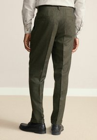Dunkelgrüne Hose mit strukturiertem Gewebe, im taillierten Schnitt, zwei Gesäßtaschen und einem umgeschlagenen Saum. Kombiniert mit schwarzen Schuhen.