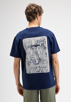 Mann, der wegschaut und ein marineblaues T-Shirt mit großem weißem Text und einer Grafik eines Hakens auf dem Rücken trägt.