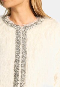 Prenda de piel sintética blanca con un cuello de cuentas y cierre frontal, adornada con piedras preciosas plateadas y transparentes dispuestas en diferentes tamaños.