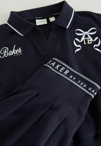 Polo azul marino con cuello, con detalles en blanco. Logo bordado "Baker" e iniciales "TB". Puños y bajo acanalados. Textura suave.
