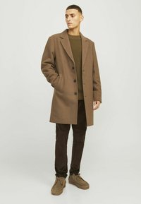 Abrigo de lana marrón con solapas muesca, cinco botones negros, usado sobre un suéter azul de punto y pantalones de pana marrones, combinado con zapatos beige.