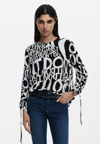 Blusa a maniche lunghe bianca e nera con un motivo a testo audace. Presenta maniche arricciate e un collo rotondo, abbinata a jeans blu scuro.
