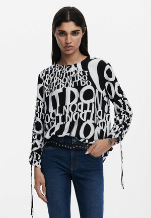 Blusa de manga larga en blanco y negro con un estampado de texto audaz. Presenta mangas con recogido y un escote redondo, combinada con jeans de color azul oscuro.