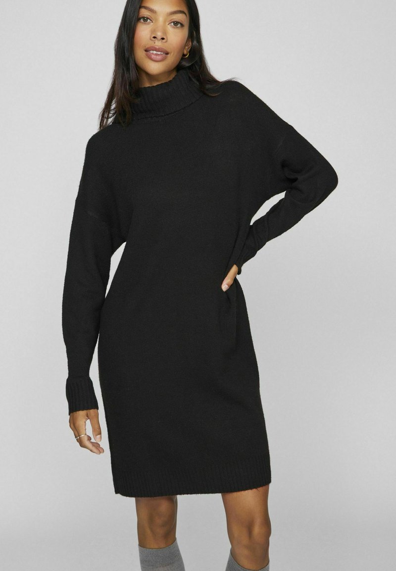 Black, knitted turtleneck dress with long sleeves and ribbed cuffs, featuring a relaxed fit and mid-thigh length.