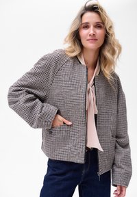 Brązowa i kremowa bomberka w wzór houndstooth z obniżonymi ramionami, ściągaczami przy mankietach i bocznymi kieszeniami, noszona na jasnej bluzce.