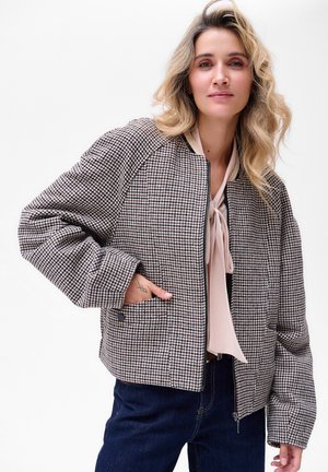 Veste bomber à motif pied-de-poule marron et crème, avec des épaules tombantes, des poignets côtelés et des poches latérales, portée par-dessus une blouse claire.