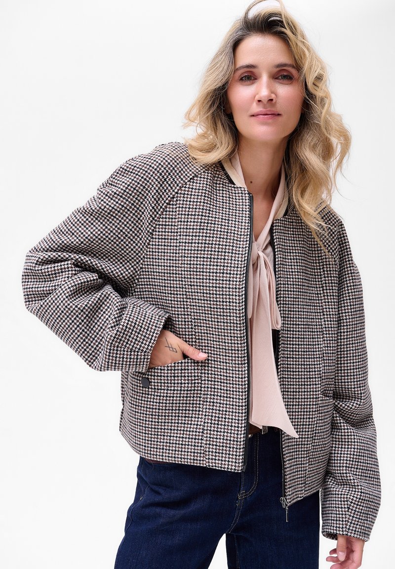 Brązowa i kremowa bomberka w wzór houndstooth z obniżonymi ramionami, ściągaczami przy mankietach i bocznymi kieszeniami, noszona na jasnej bluzce.