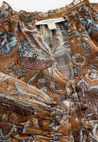 Hnedá kvetinová blúzka so zberaným výstrihom, textúrovanou tkaninou a metalickými vláknami, s modro-bielymi kvetinovými vzormi a gombíkmi.