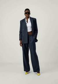 Mornarsko prugast suit s dvostrukim krojem, širokimi hlačami in belo skrajšano srajco, v kombinaciji z rumenimi špičastimi petami in sončnimi očali.