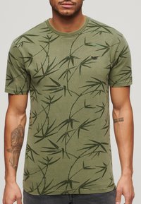 Camiseta de algodón verde oliva con un patrón de hojas de bambú, diseño de cuello redondo y un pequeño logo de marca en el pecho.