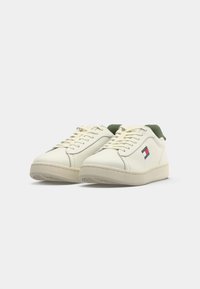 White leather sneakers with green accents, round toe, textured sole, and laces. Features a small logo patch on the side.