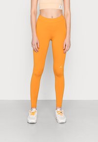 Leggings arancioni con un tessuto liscio ed elasticizzato. Presentano un alto punto vita e un piccolo logo bianco sulla parte inferiore della gamba. Indossati con sneaker bianche.