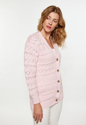 Frau trägt eine hellrosa gestrickte Strickjacke mit Knopfleiste vorne und weiße Hose, schaut leicht nach links vor weißem Hintergrund.
