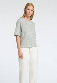Maglione grigio chiaro a maniche corte realizzato in materiale morbido, con collo rotondo e cinque bottoni. Abbinato a pantaloni di colore chiaro, con vestibilità ampia.