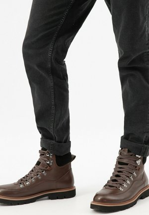 Une personne portant un jean noir roulé et des bottes de randonnée en cuir marron avec des œillets en métal sur fond blanc.