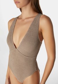 Ruskea neuleuima-asu, jossa on syvä V-pääntie ja leveät hihnat. Asussa on teksturoitu kuvio ja se on muotoiltu tiukasti.