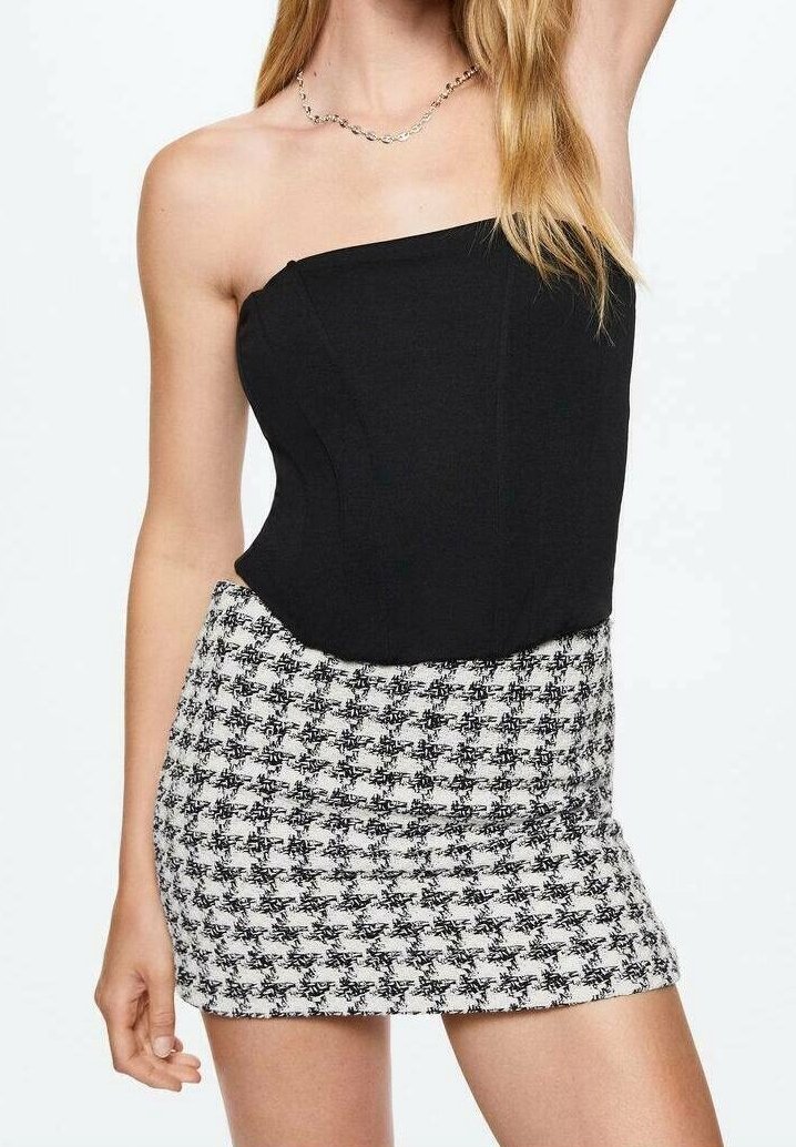 Femme portant un top noir à une épaule et une mini-jupe à taille haute pied-de-poule noire et blanche, avec de longs cheveux blonds et un collier chaîne.