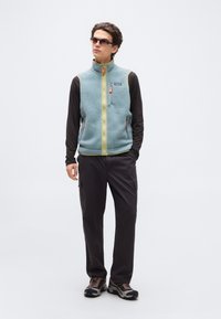 Άνδρας που φοράει μπλε φλις γιλέκο με φερμουάρ πάνω από μαύρο μακρυμάνικο πουκάμισο, μαύρο παντελόνι cargo, παπούτσια πεζοπορίας και γυαλιά ηλίου, στέκεται μπροστά σε λευκό φόντο.