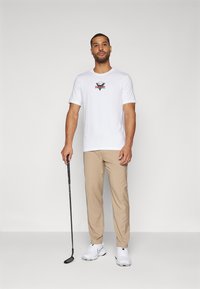 Balta trumpa rankovėmis marškinėliai su įvairiaspalviu Nike logotipu, derinami su smėlio spalvos kelnėmis ir baltomis sportinėmis bateliais, laikantys golfo lazdelę.
