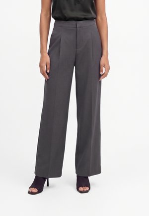 Femme portant un pantalon gris taille haute à jambes larges associé à des bottines noires à talons hauts et bouts ouverts sur fond blanc.