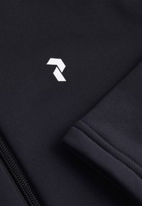 Sort materiale med en struktureret finish, der har et hvidt geometrisk logo og en lynlåslukning. Designet inkluderer en ribbet manchet-detail.