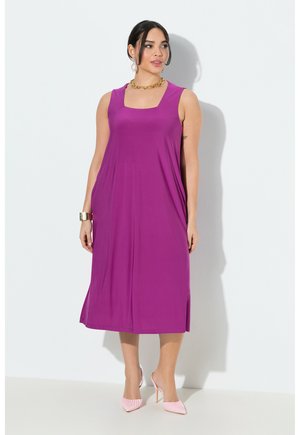 Kvinde iført en ærmeløs lilla midi-kjole med firkantet udskæring, guldkæde, armbånd, øreringe og pink højhælede sko.