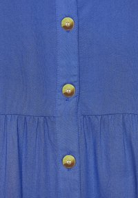 Blauer Stoff mit glatter Textur, der eine Reihe von fünf runden, glänzenden goldenen Knöpfen und gerafftem Stoff unterhalb der Knöpfe aufweist.