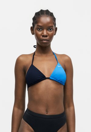 Femme aux cheveux tressés portant un haut de bikini triangle bicolore avec un bas noir, debout devant un fond clair uni.