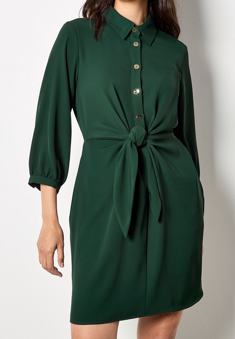 Femme portant une robe boutonnée vert foncé avec des manches trois-quarts et un détail de nœud noué à la taille, debout contre un mur blanc.