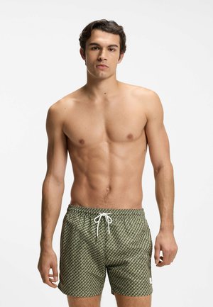 Jonge man met kort donker haar die groene zwemshorts met patroon en witte trekkoord draagt, staand tegen een witte achtergrond.
