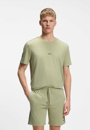 Junger Mann trägt hellgrünes BOSS-T-Shirt und passende Shorts, steht mit einer Hand in der Tasche vor weißem Hintergrund.