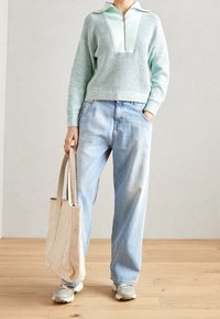 Hellgrüner Strickpullover mit Reißverschlusskragen, kombiniert mit lockeren hellblauen Jeans. Trägt eine beige Tote-Bag und graue Sneakers.