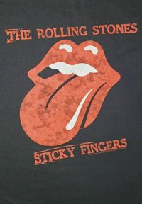 Svart T-skjorte med en stor rød Rolling Stones tungelogo med en hvit munn, samt teksten "THE ROLLING STONES" og "STICKY FINGERS" i oransje.